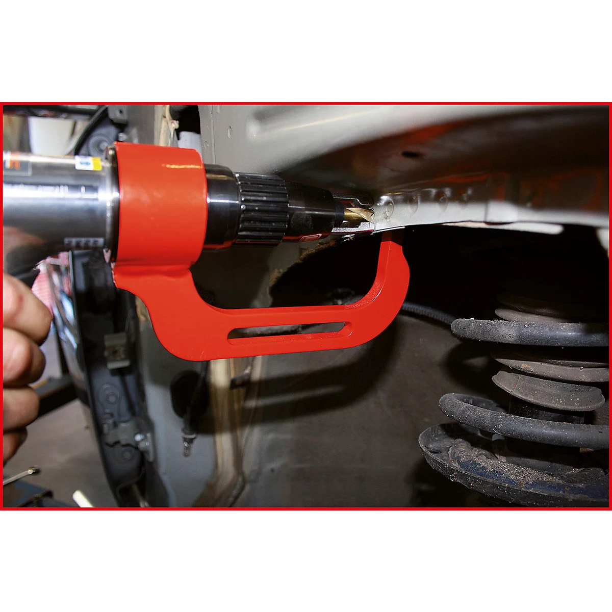 Eine Person verwendet ein rotes Werkzeug, um ein Bauteil in einem Auto zu bearbeiten. Im Hintergrund ist eine Schraubenfeder zu sehen.